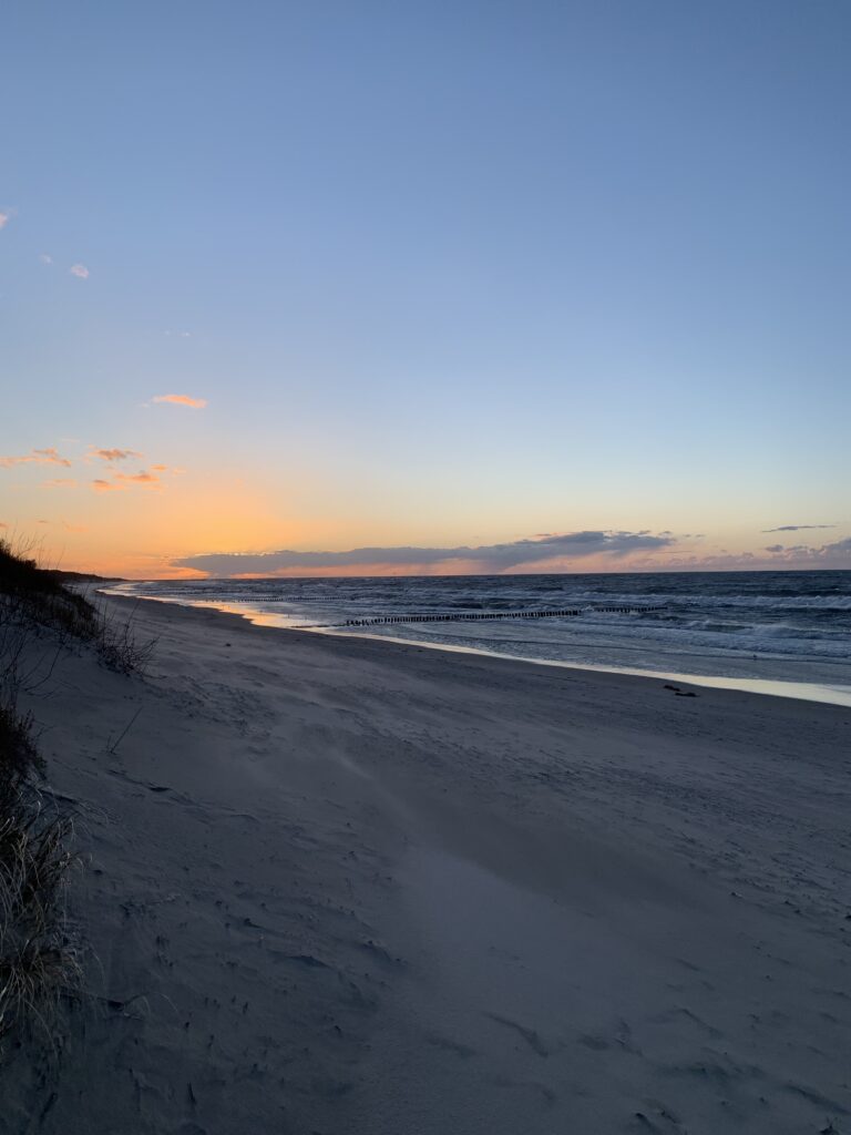 Das Bild ist Richtung Westen fotografiert. Es ist ein langer, leerer Strand zu sehen. Im Hintergrund ist die Sonne bereits untergegangen und hinterlässt orange-rote Farben am Himmel.